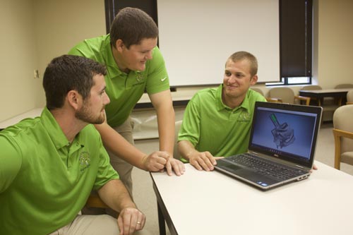 Three young men in green polo shirts gathered around a laptop in a classroom