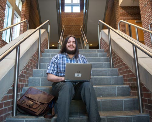 A young bearded man sitting on a staircase with an open laptop on his lap