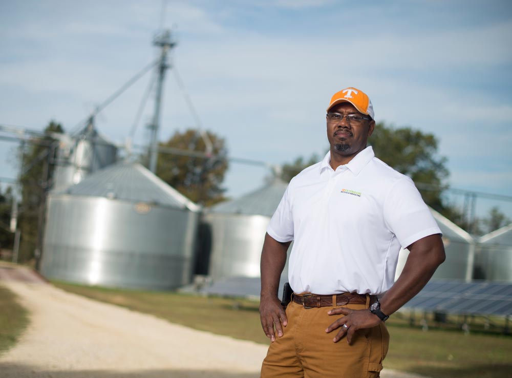 JC Dupree stands in front of agricultural buildings waring a Volunterrs baseball cap and work wear