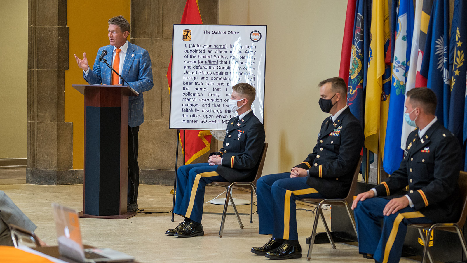 Randy Boyd speaks at a ROTC commissioning ceremony, with 3 student cadets seated in front of service flags